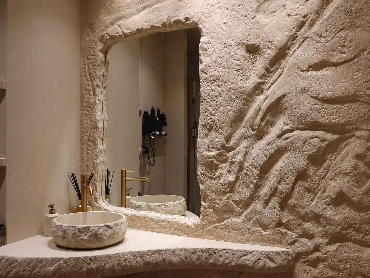 Aseo de Nudhe Studio con lavabo de piedra, grifería dorada y pared esculpida a mano